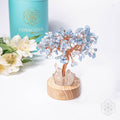 Decorative crystal tree on a wooden base with flowers and a cylindrical container in the background.